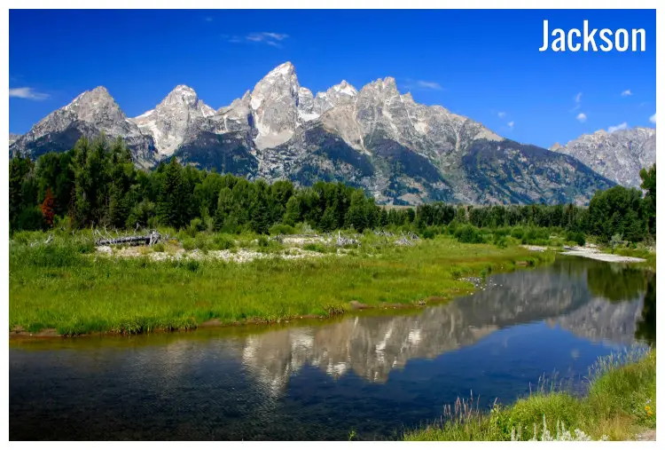 A calm and clear river and vast green fields approaching the iconic white mountains within the Grand Teton National Park in Jackson, Wyoming