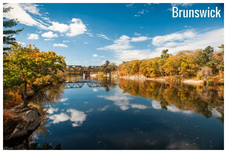 The calm Androscoggin River in Brunswick, Maine, with a rail bridge on it and scenic brown and golden trees on both sides mirrored in the water The calm Androscoggin River in Brunswick, Maine, with a rail bridge on it and scenic brown and golden trees on both sides mirrored in the water