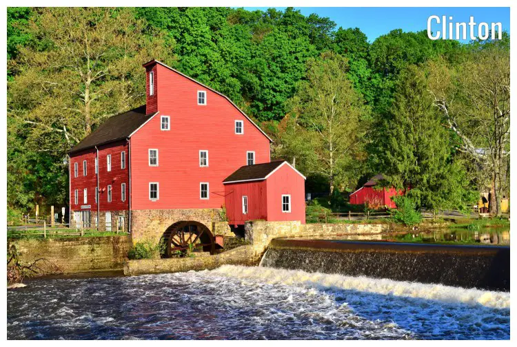 A bright red house next to a dense tree forest, with a mill and dam water flowing over in a rural area in Clinton, New Jersey